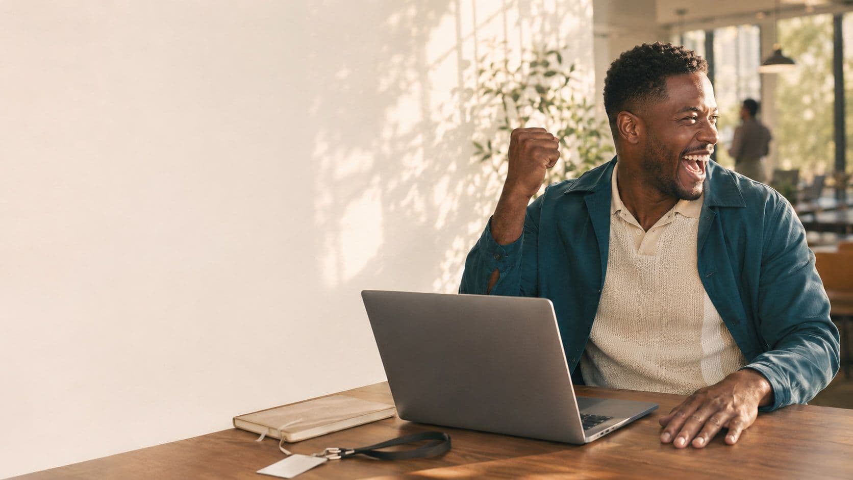 A man at a laptop punching the air and grinning, alone at a sunlit table